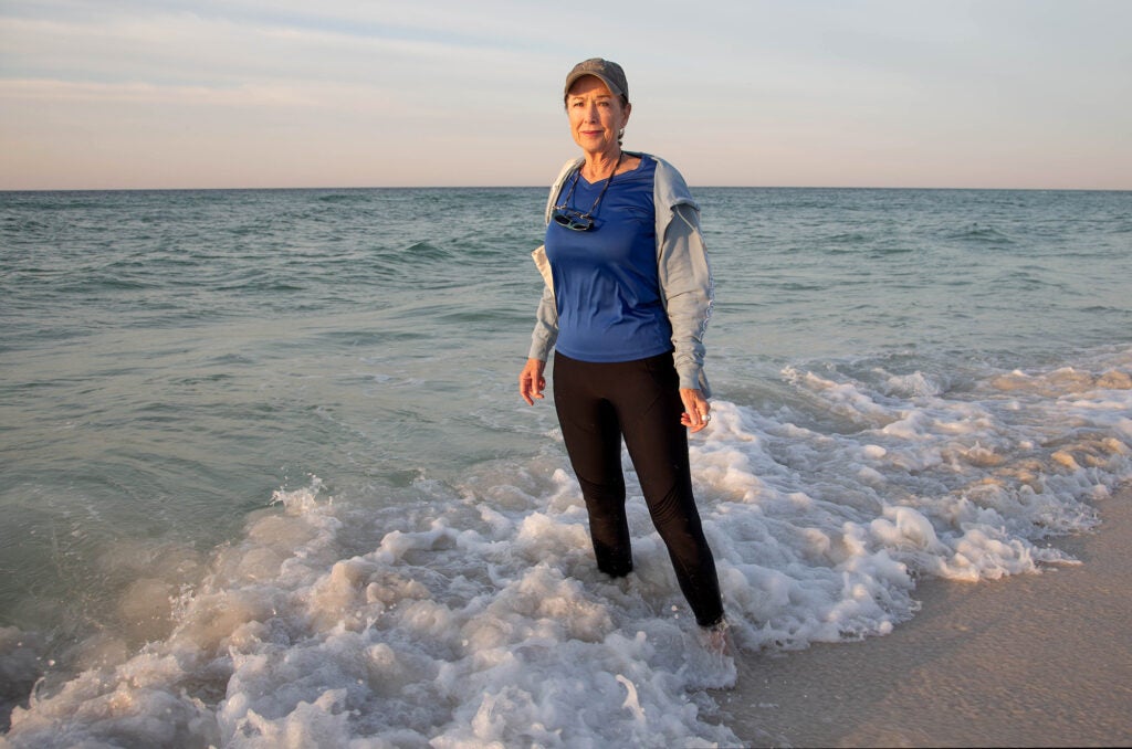 A woman wearing a hat, sweatshirt and black pants stands in ankle-deep water with the ocean behind her, looking directly at the camera with a serious expression.