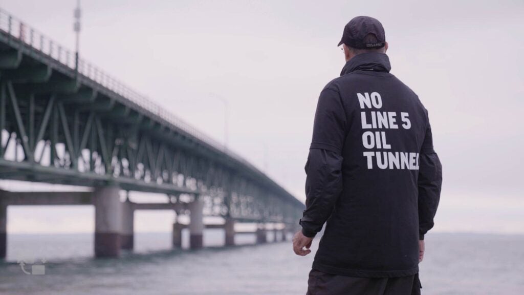 A man wearing a sweatshirt that says "No Line 5 Oil Tunnel" facing a body of water and a bridge